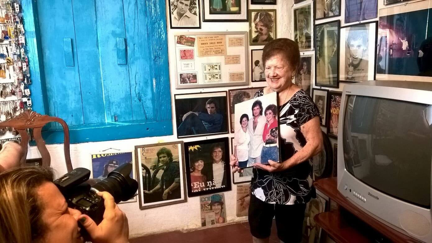 A imagem mostra Gercy Volpato sendo fotografada na sala de sua casa, dedicada ao acervo do cantor Roberto Carlos.