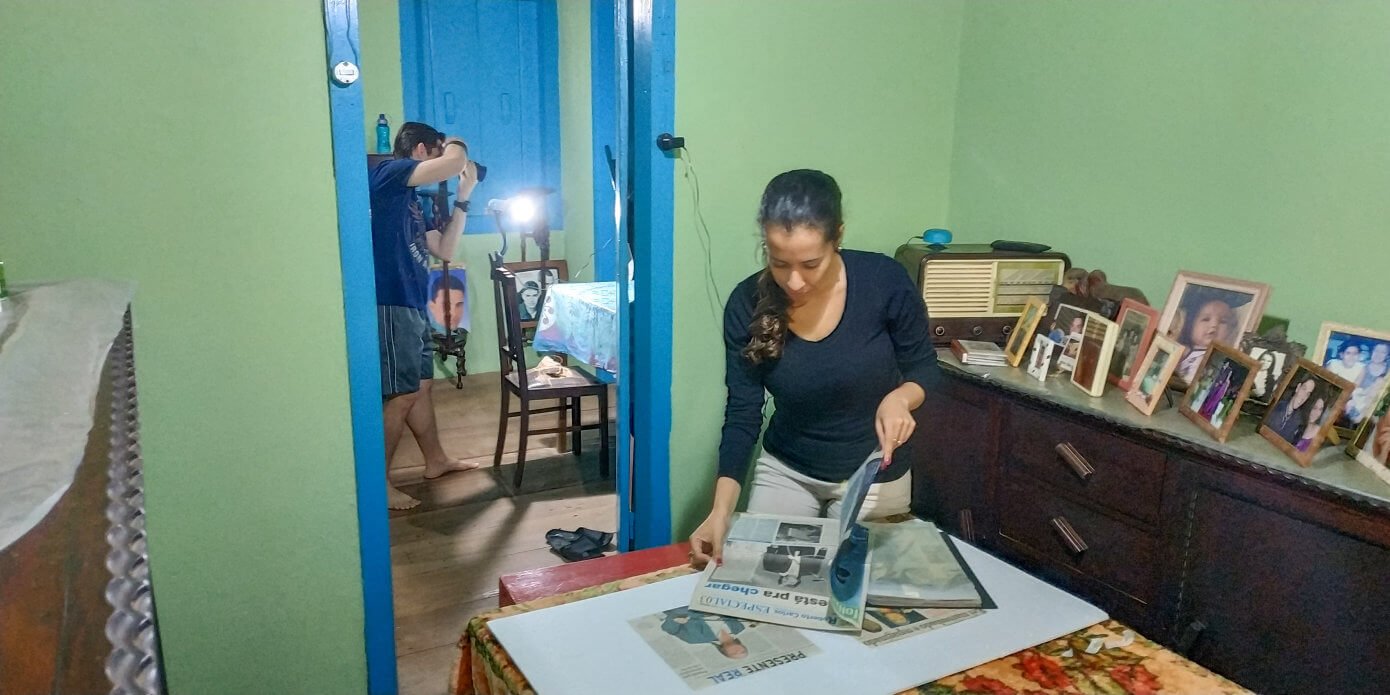 Luan Volpato fotografando recortes de jornal enquanto Sara Moreira folheia jornais para encontrar imagens e notícias de Roberto Carlos para serem fotografadas.